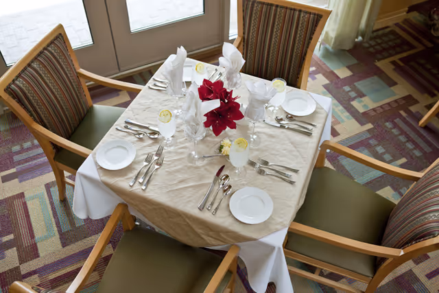 Overhead view of a set dining table for four with folded white napkins, glassware with lemon slices, a red floral centerpiece, and four chairs on patterned carpet near glass doors.