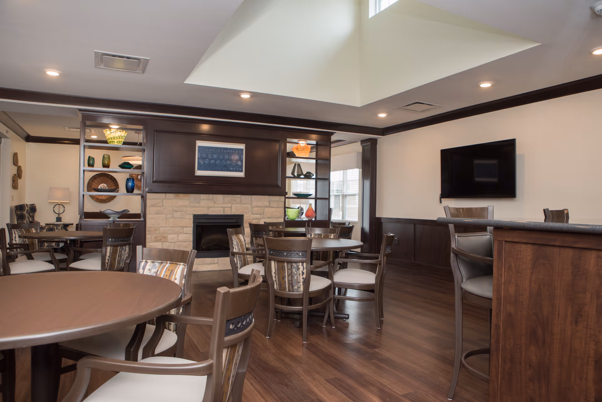 A cozy interior common area with multiple round wooden tables and chairs arranged on a polished wooden floor. The room features a stone fireplace with a dark wood mantle, built-in shelves displaying decorative items, a wall-mounted flat-screen TV, and a bar counter with high chairs. The ceiling has recessed lighting and a skylight allowing natural light to brighten the space.