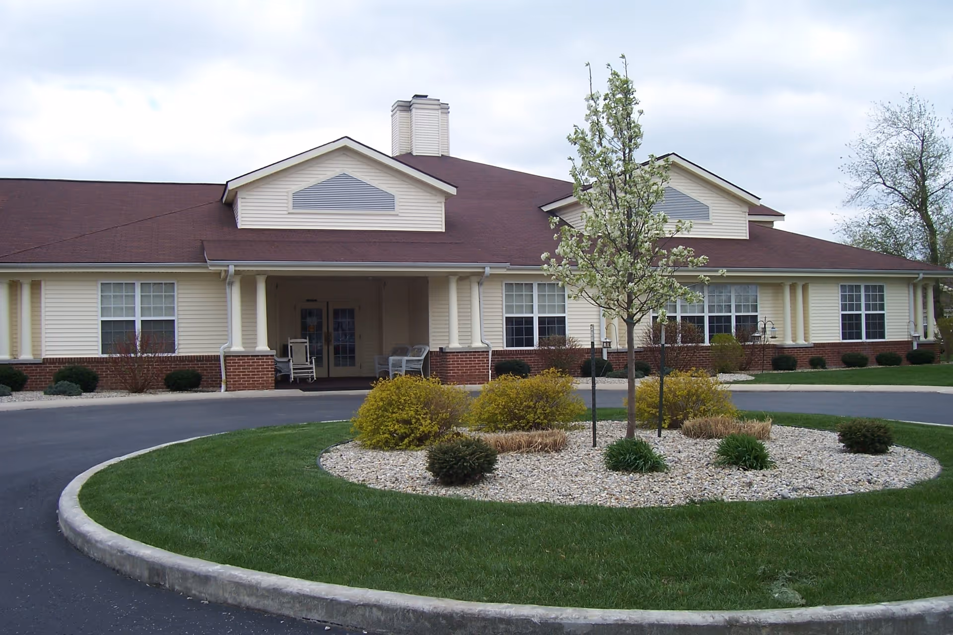 Front exterior view of a single-story building with beige siding and a brown roof, featuring a covered entrance with chairs on the porch. In front of the building is a circular landscaped area with a small tree and shrubs surrounded by grass and a paved driveway.