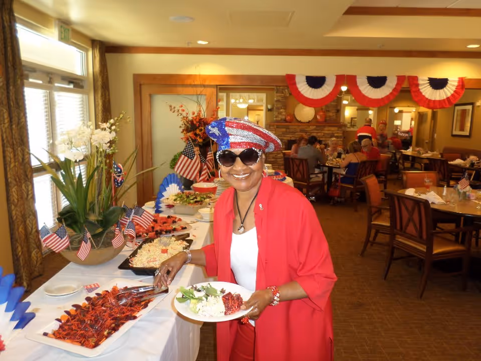 A woman wearing a red, white, and blue sequined hat and sunglasses is serving herself food from a buffet table decorated with small American flags. The room is decorated with red, white, and blue bunting, and several people are seated at tables in the background, dining and socializing.