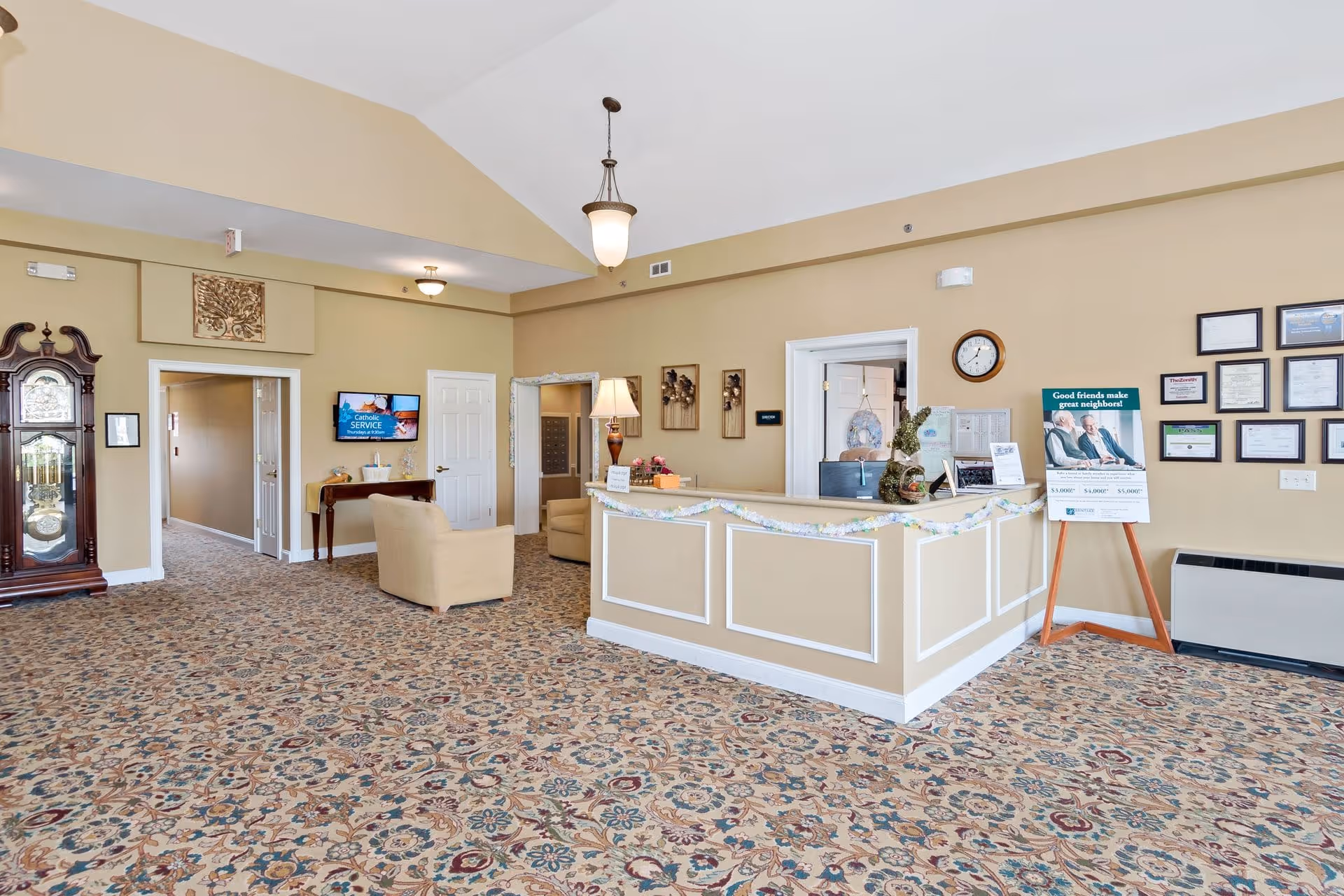 Reception area of Bentley Assisted Living at Branchville with a beige front desk decorated with garland, a patterned carpet, beige walls, a grandfather clock, two beige armchairs, a wall-mounted TV displaying a Catholic service announcement, framed certificates on the wall, and a sign on an easel that reads 'Good friends make great neighbors!'
