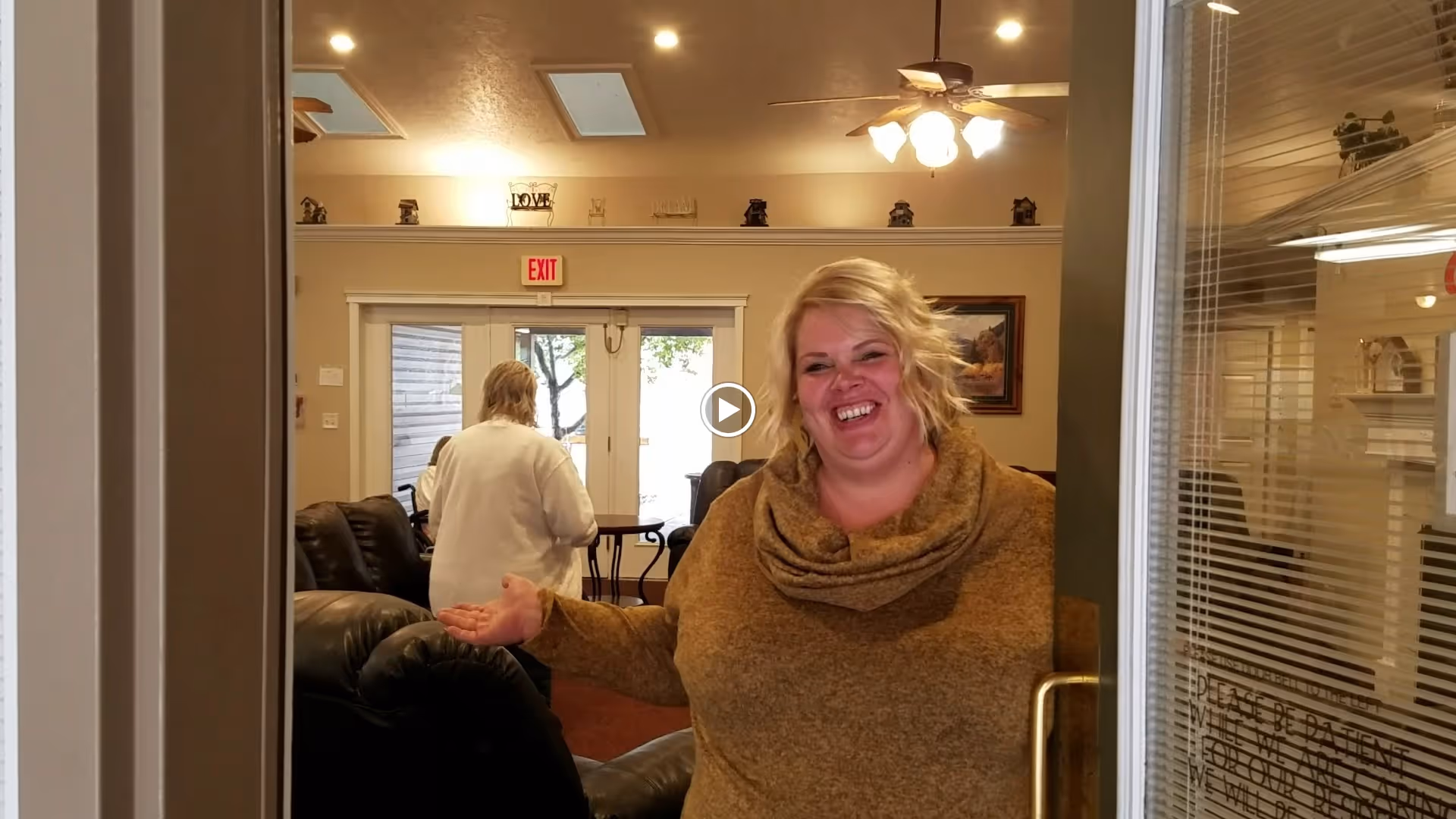 A smiling woman stands in a doorway gesturing into a cozy common room with recliners and another person in the background.