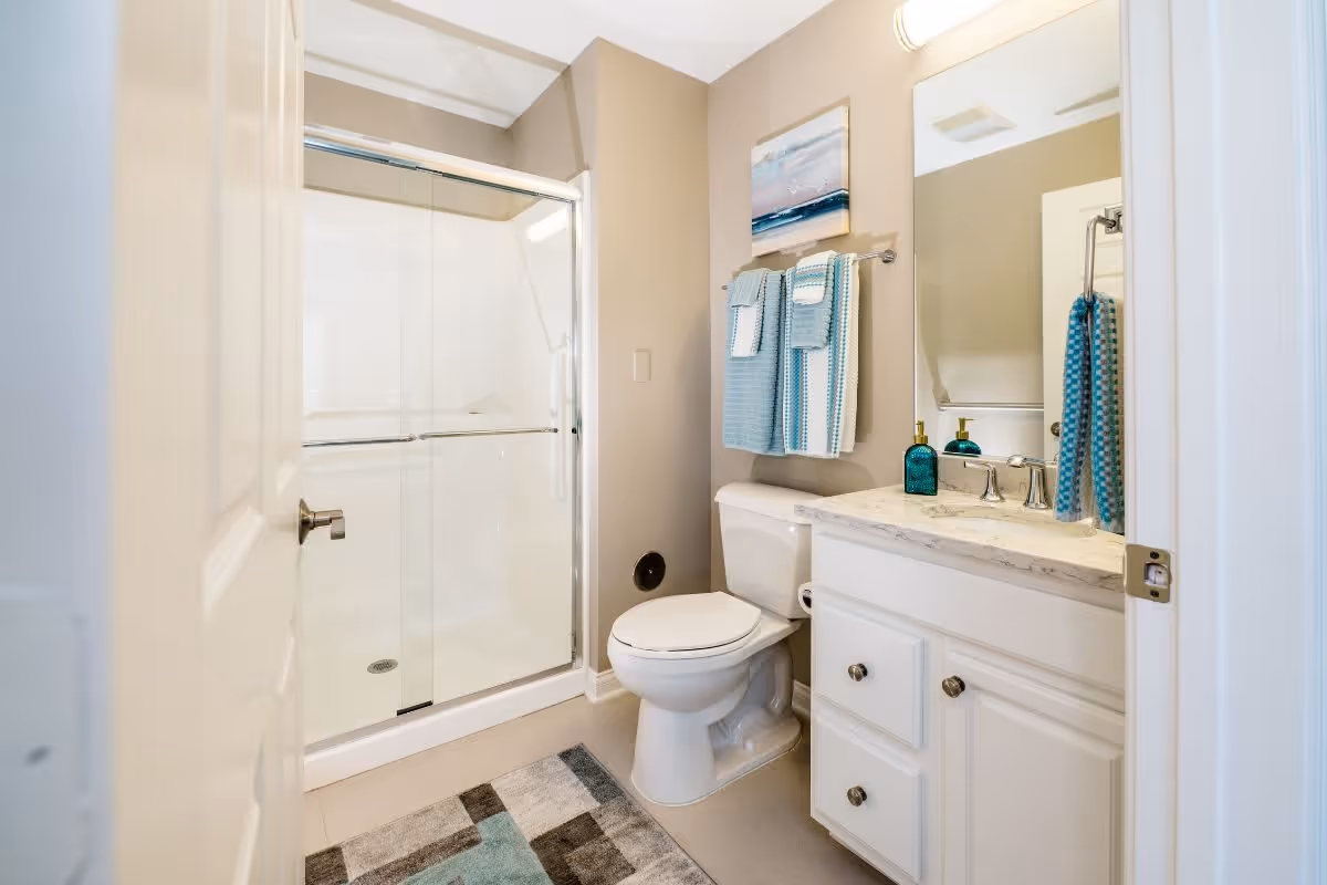 Bright bathroom with a glass-enclosed shower, white toilet and vanity with marble countertop, mirror, and teal towels.