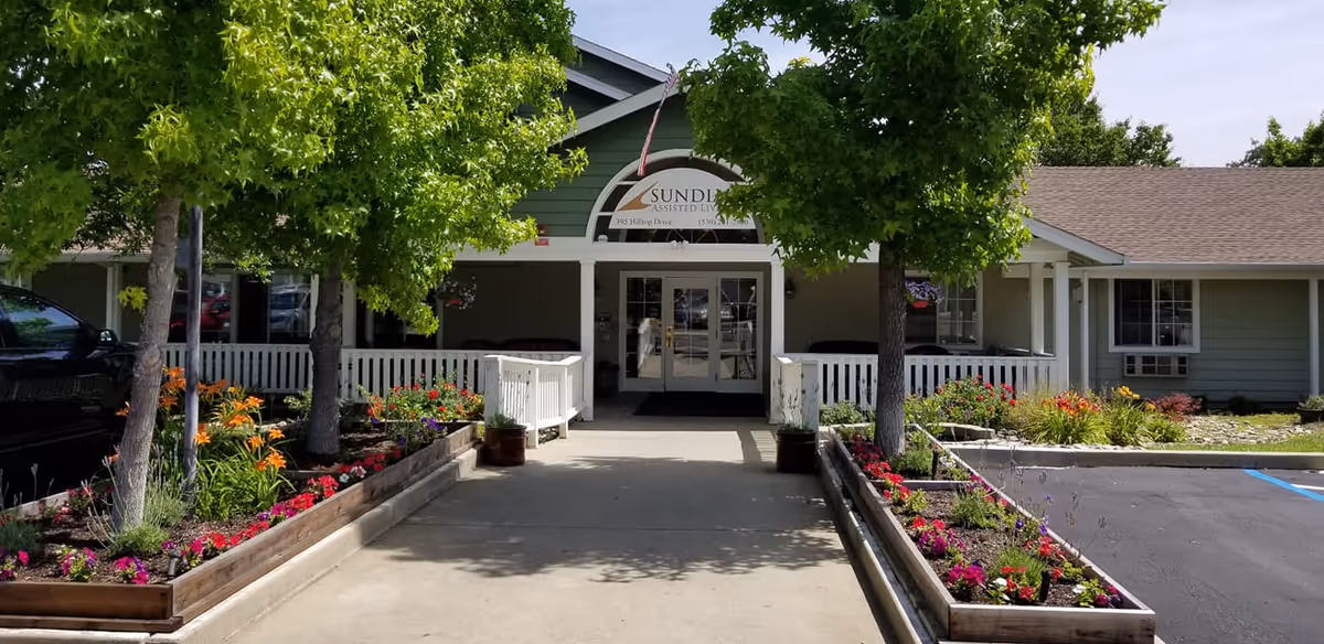 Entrance to Sundial Assisted Living facility with a concrete walkway flanked by flower beds and trees, leading to double glass doors under a green gabled roof with a white picket railing on the porch.