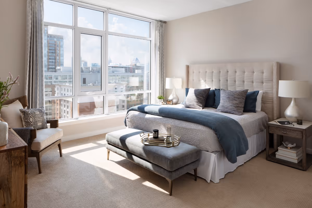 Bright bedroom with a large window showing a cityscape view. The room features a tufted headboard bed with blue and gray pillows and a blue throw blanket. A gray upholstered bench sits at the foot of the bed with a tray on top. There are two bedside tables with lamps, a wooden dresser, and an armchair with cushions. The room is carpeted and decorated in neutral tones.
