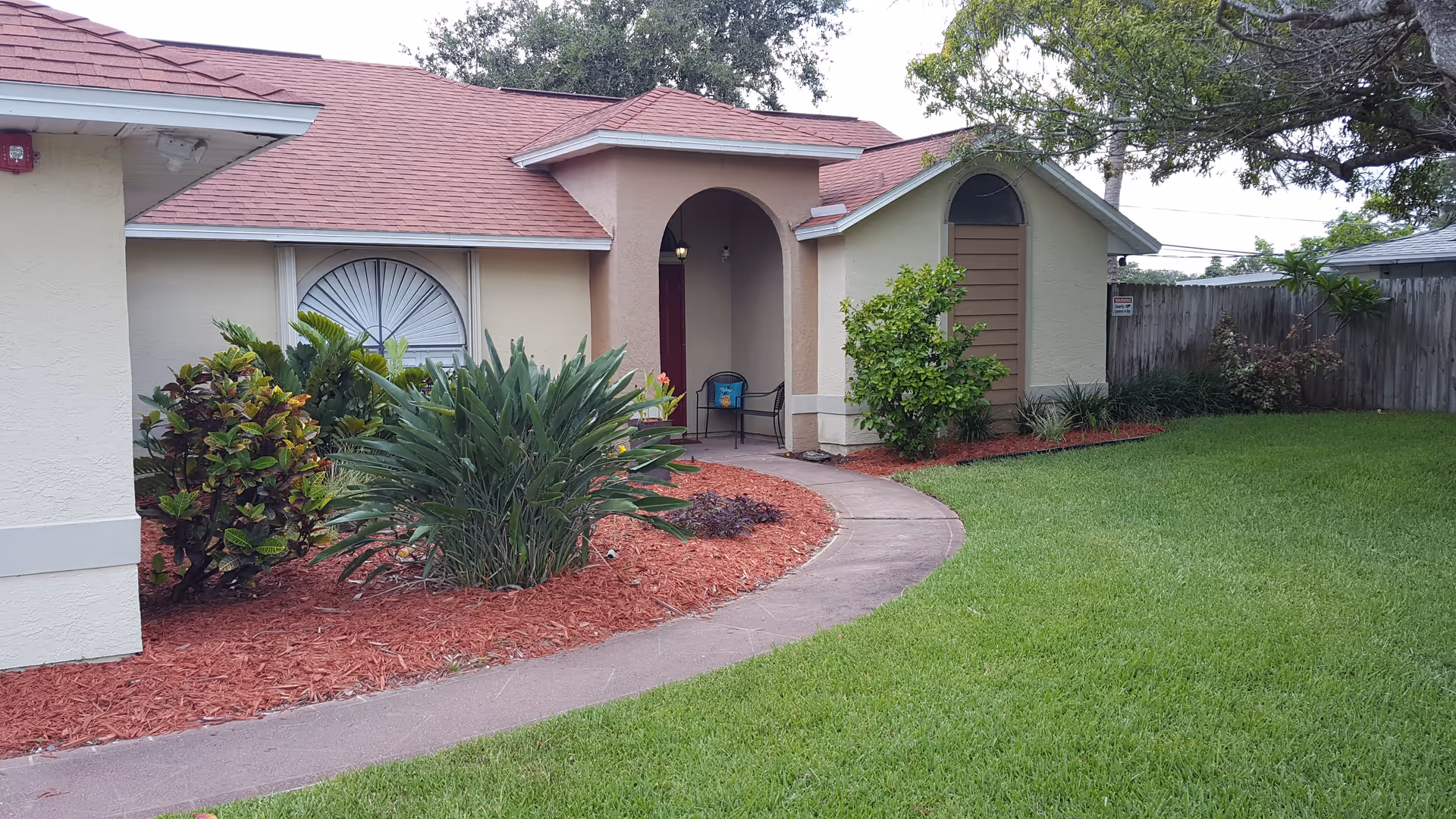 Front entrance of a single-story house with a curved walkway, green lawn, and landscaped beds.