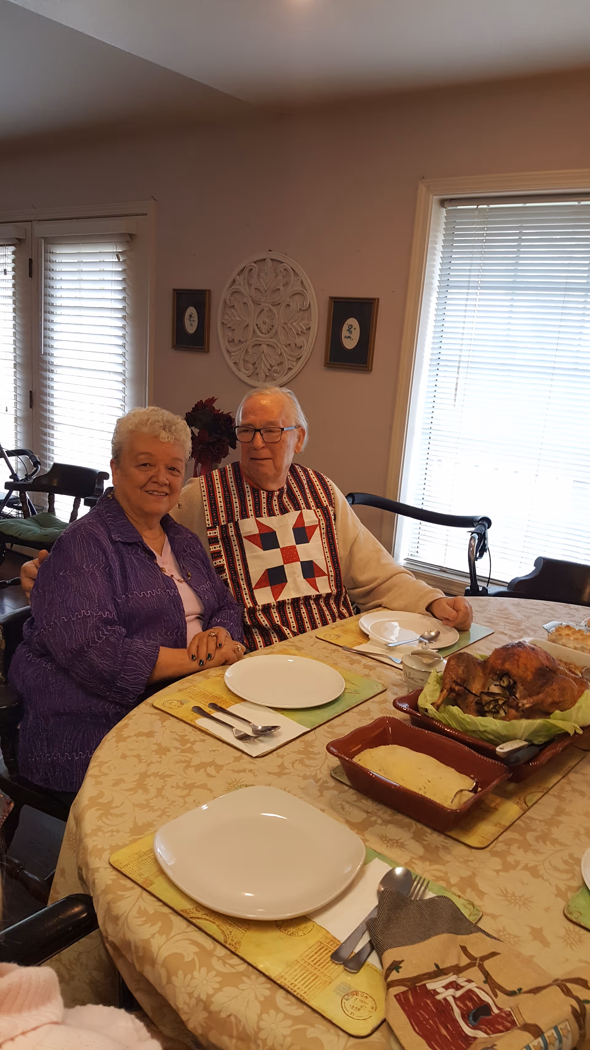 An elderly couple sitting at a dining table set with plates, utensils, and food including a roasted turkey and a casserole dish. The room has windows with blinds and decorative wall art behind them.