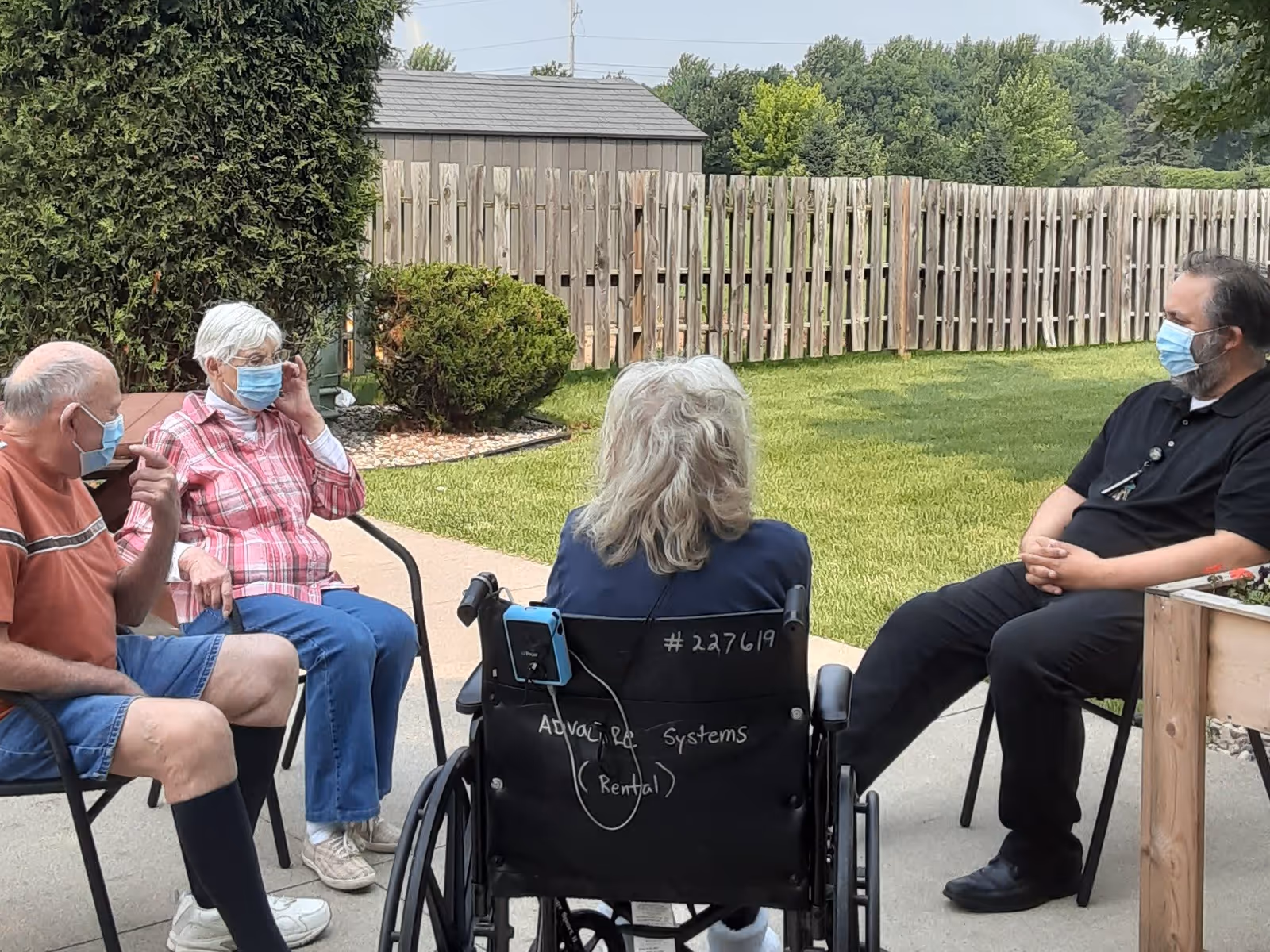 Four people sitting outside in a garden area with a wooden fence and greenery in the background. Three elderly individuals, two women and one man, are seated on chairs and a wheelchair, all wearing face masks. A man in black clothing, also wearing a face mask, is seated opposite them, engaged in conversation.