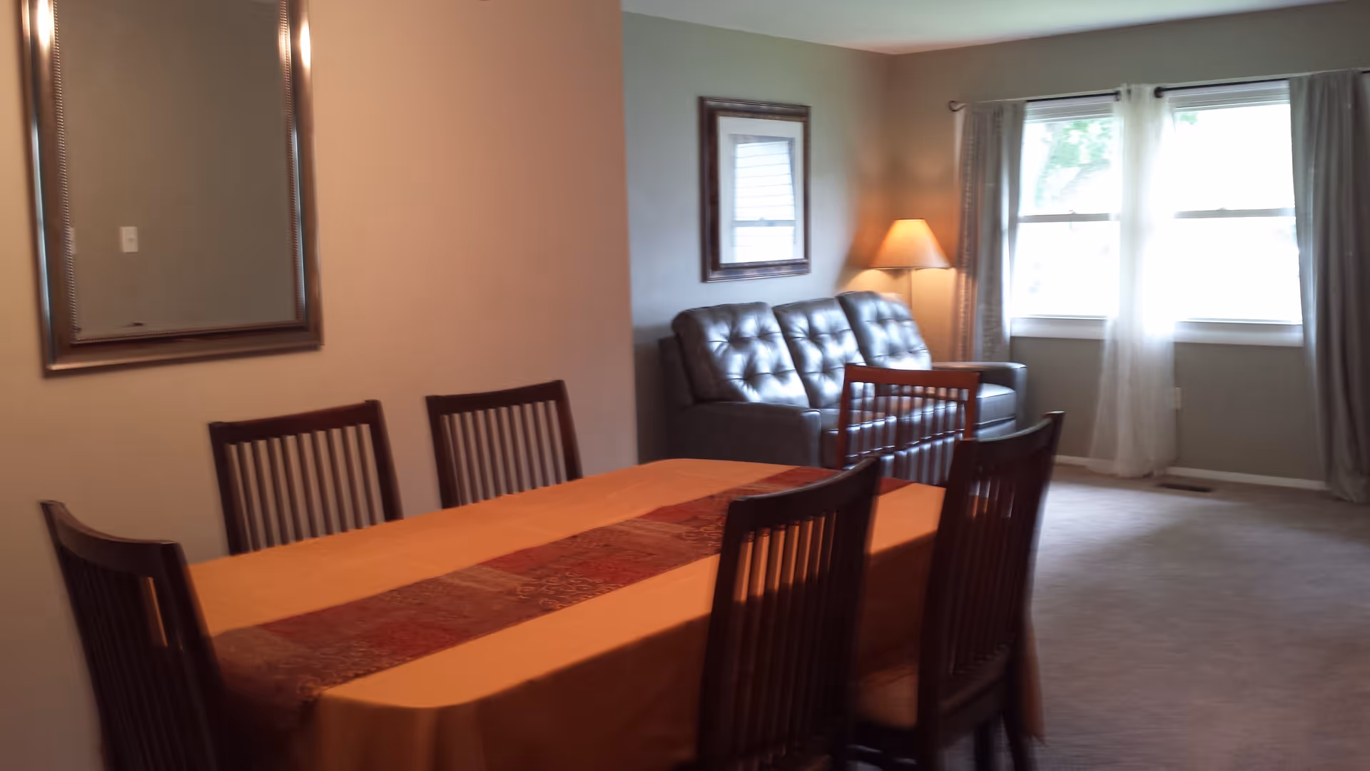 A dining area with a rectangular table covered with an orange tablecloth and a decorative runner, surrounded by six wooden chairs. In the background, there is a gray leather sofa, a floor lamp with a beige shade, a framed picture on the wall, and two windows with sheer white curtains allowing natural light to enter.