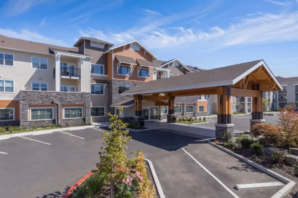 Exterior view of The Springs at Sherwood senior living facility showing a multi-story building with stone and wood accents, a covered entrance with wooden beams, a parking area, and landscaped greenery under a blue sky.