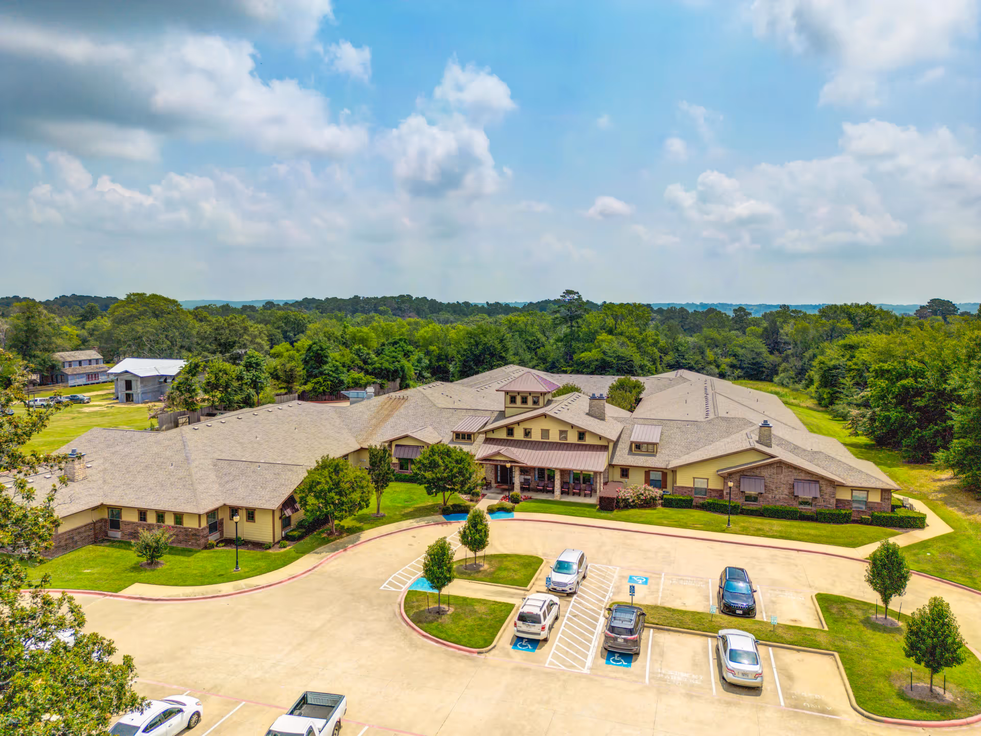 Aerial view of the assisted living facility building, parking lot, and surrounding trees under a partly cloudy sky.