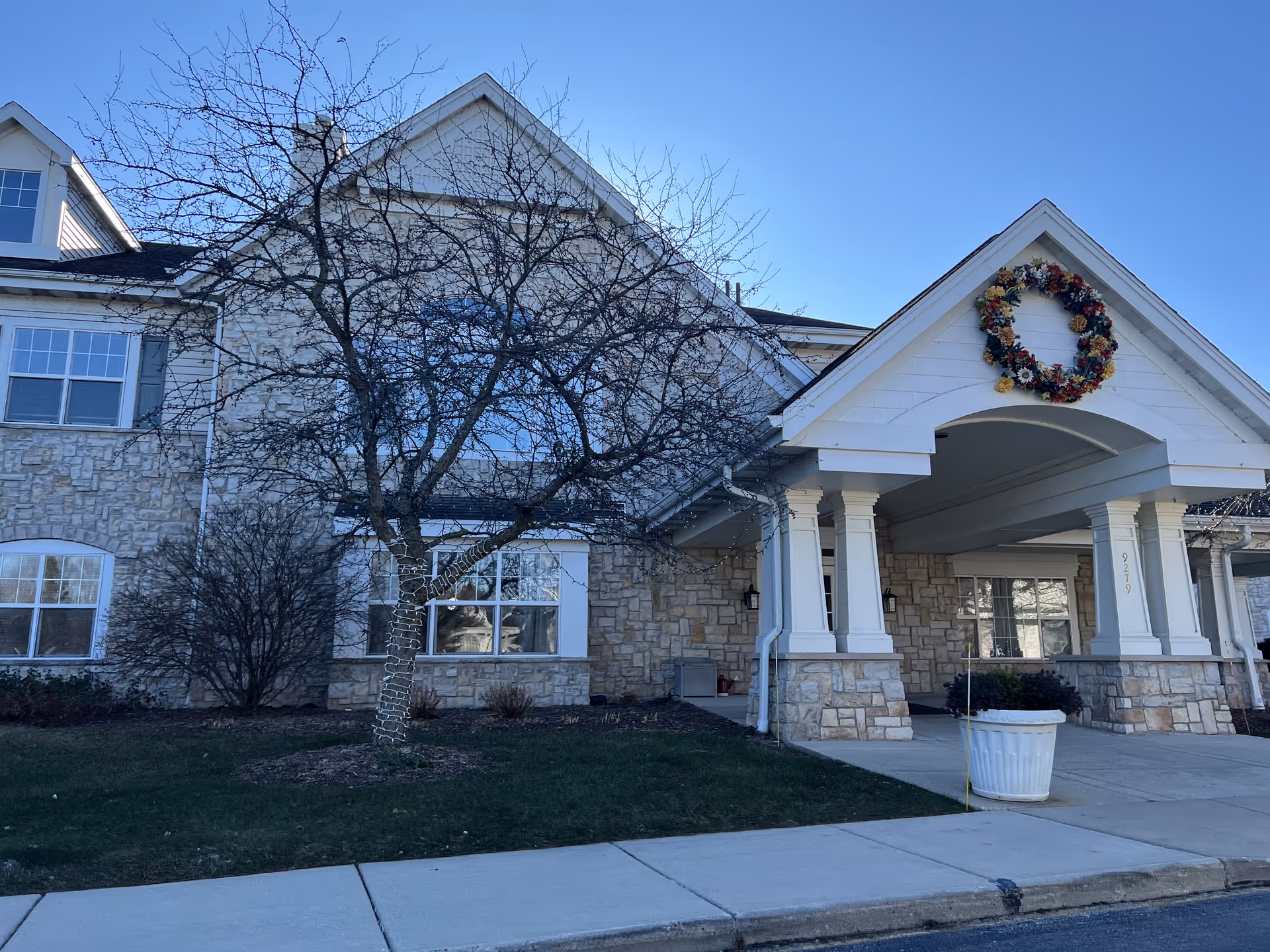 Exterior view of a senior living facility building with stone and white siding, a covered entrance decorated with a large floral wreath, leafless trees, and a clear blue sky.