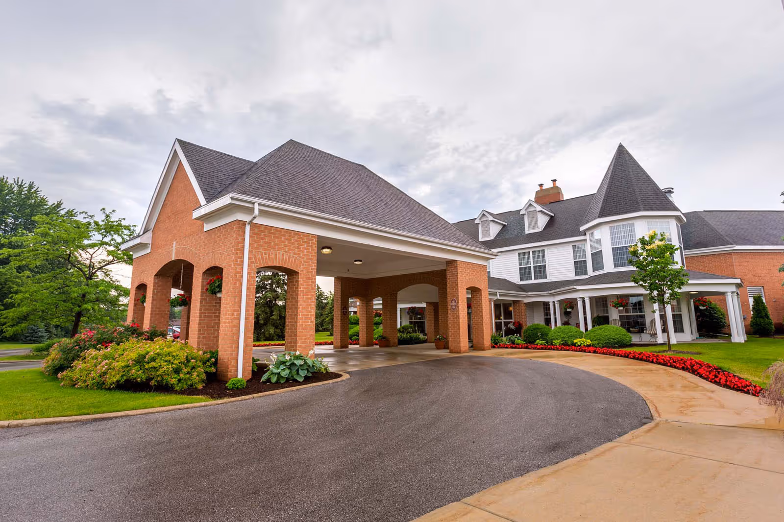 Exterior view of a senior living facility with a covered driveway entrance, brick pillars, well-maintained landscaping with green bushes and red flowers, and a large building with white siding and multiple windows under a cloudy sky.