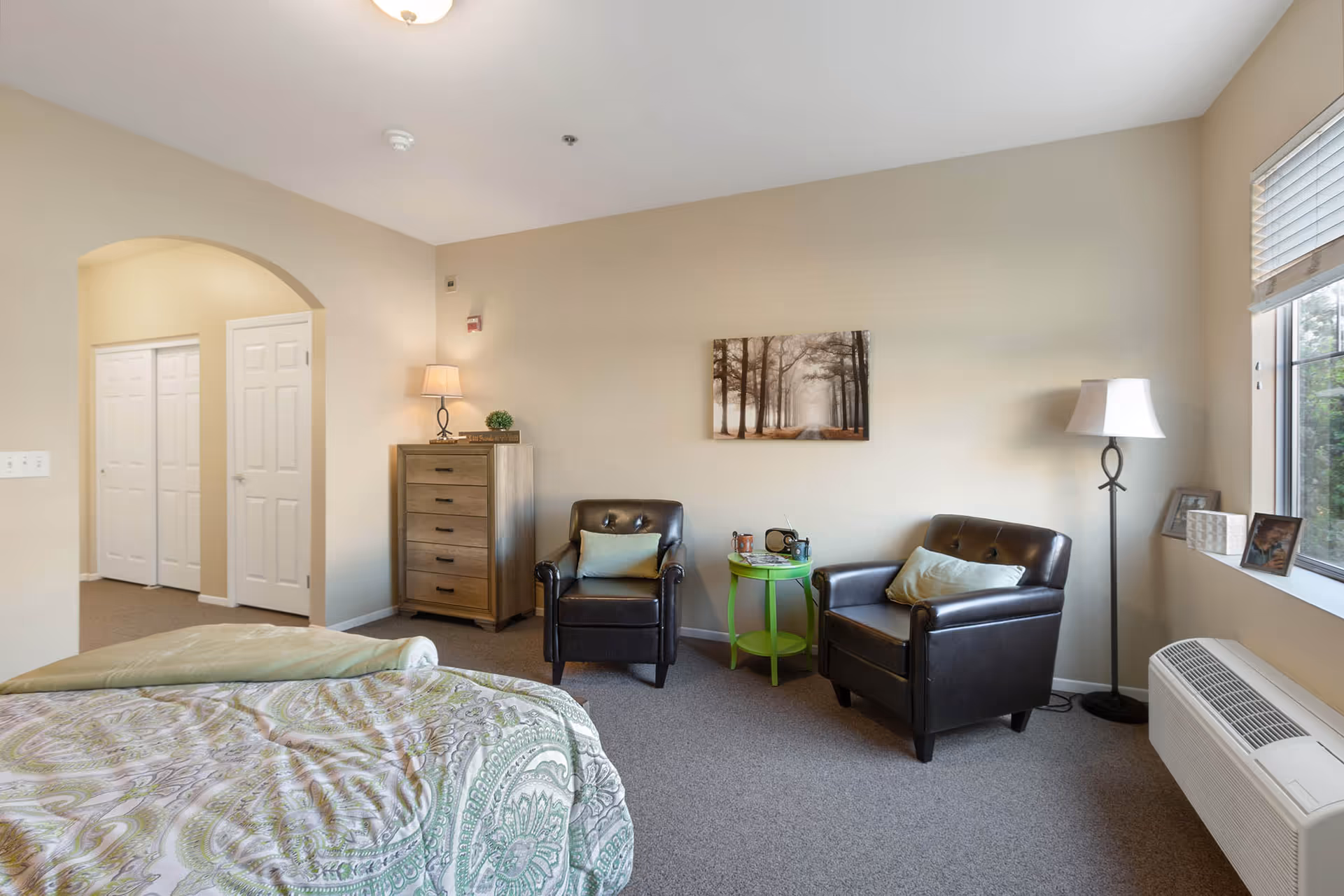 A cozy bedroom with a bed covered in a green and white patterned comforter. Two dark brown leather armchairs with light green pillows are positioned around a small green side table holding a radio and a cup. A wooden chest of drawers with a lamp and small plant sits against the wall. A floor lamp stands near a window with blinds, and framed photos are placed on the windowsill. The walls are painted beige, and a picture of a tree-lined path hangs above the chairs.