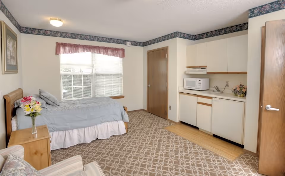 A small studio room with a single bed covered in a light blue comforter, a wooden nightstand with a vase of flowers, a window with blinds and a pink valance, a kitchenette with white cabinets, a microwave, a sink, and a dishwasher, and a patterned carpet floor. There is a wooden door and a floral border near the ceiling.
