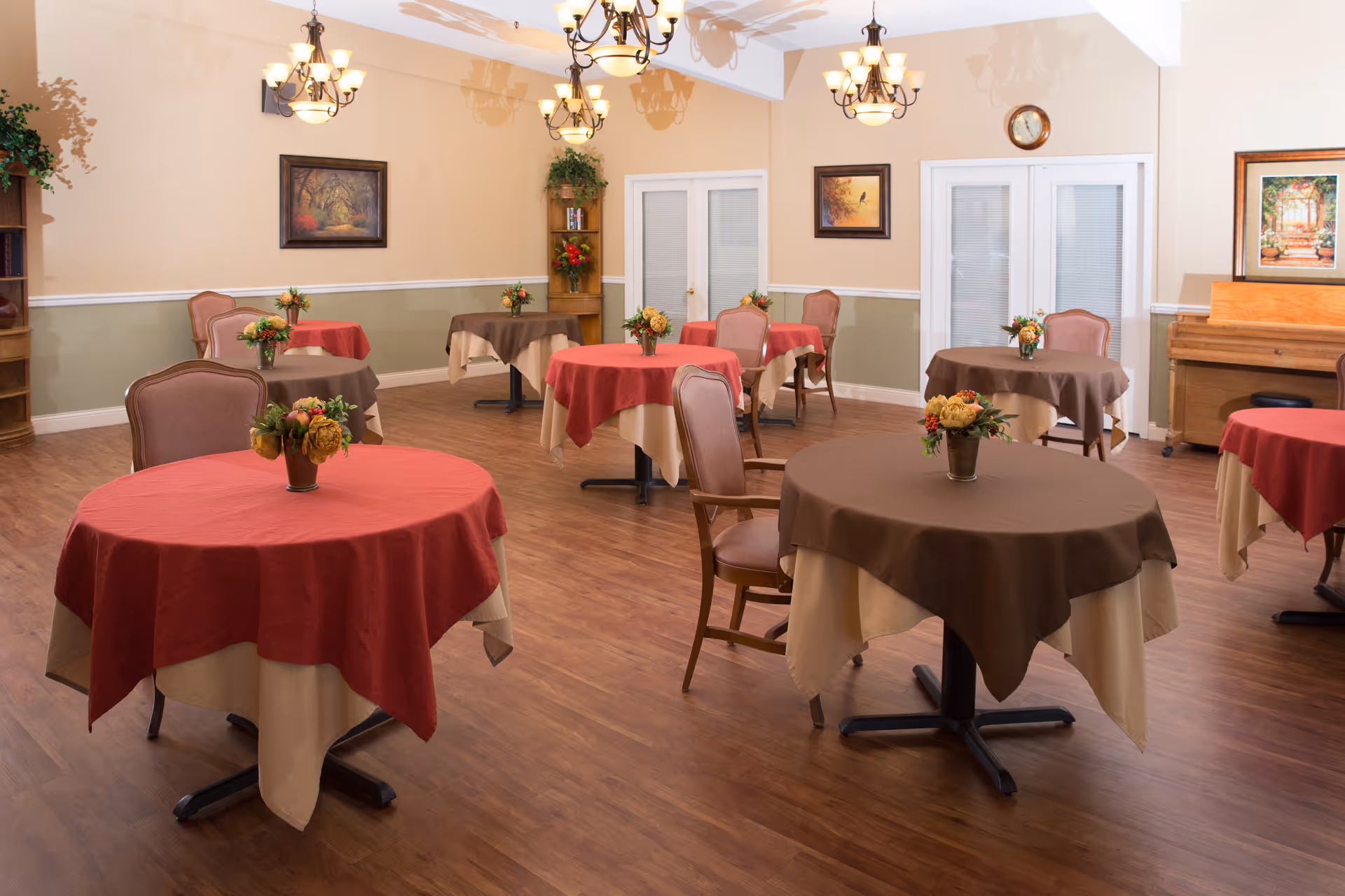 A dining room with several round tables covered with red and brown tablecloths, each adorned with a small floral centerpiece. The room has wooden flooring, beige and green walls, framed artwork, chandeliers, and a piano in the corner.