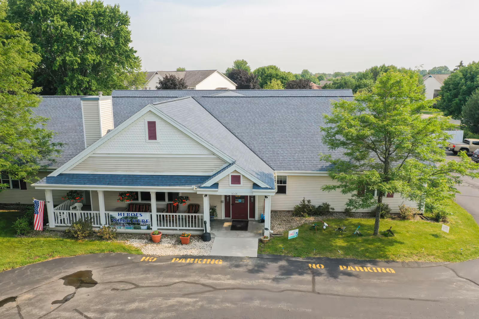 Front exterior of a single-story assisted living building with a covered porch, hanging flower baskets, an American flag, and a 'Heroes work here' banner.