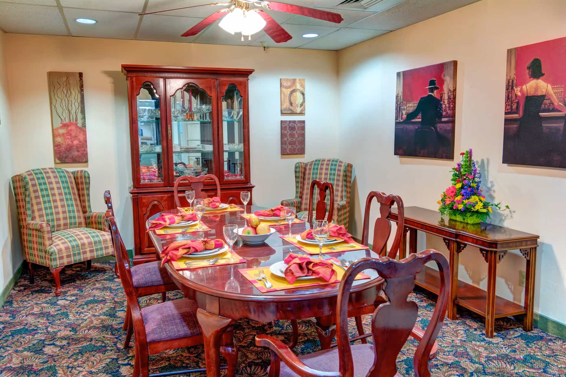A dining room with a polished wooden dining table set for six people with plates, glasses, and red napkins. The room features two plaid upholstered armchairs, a wooden china cabinet with glassware, a floral arrangement on a side table, and three paintings on the walls. A ceiling fan with red blades is visible above.