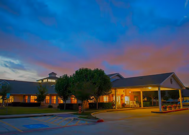 Exterior view of The Courtyards at Pasadena senior living facility at dusk, showing a single-story building with warm interior lights, a covered entrance with rocking chairs, trees, and a parking area with a handicap parking space in the foreground.
