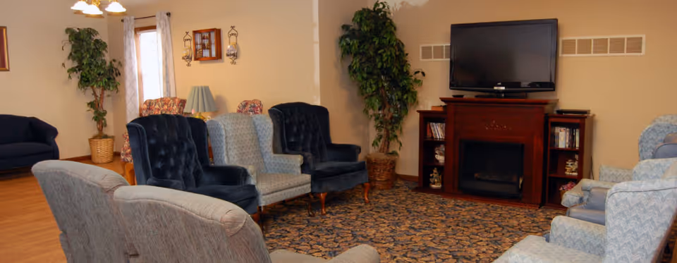 A cozy living room area in an assisted living facility featuring multiple upholstered armchairs arranged in a circle on a patterned carpet. There is a wooden fireplace with a flat-screen TV mounted above it, flanked by small shelves with decorative items. Potted plants and wall decorations add to the warm ambiance.