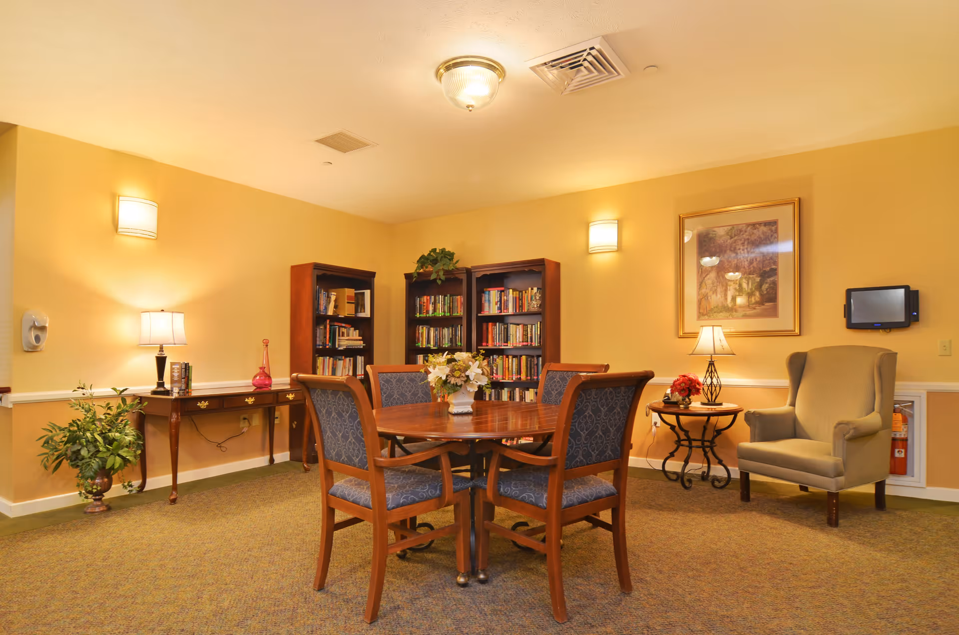 A cozy reading and sitting area with a round wooden table surrounded by four upholstered chairs. Behind the table are three wooden bookshelves filled with books. To the left, there is a wooden console table with a lamp, books, and a decorative vase, along with a potted plant on the floor. On the right side, there is a comfortable armchair next to a small round table with a lamp and a flower arrangement. The walls are painted a warm yellow, and the room is softly lit by wall sconces and ceiling lights.