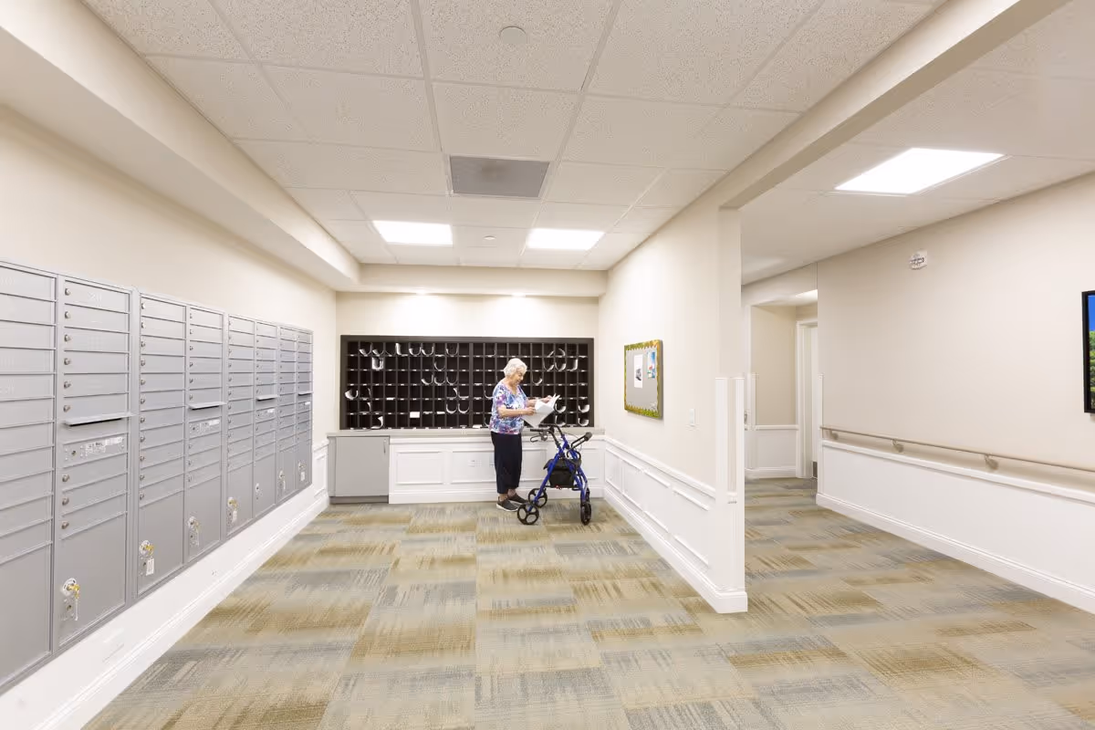 A senior woman using a walker is checking her mail in a mailroom with multiple mailboxes and cubbyholes. The room has beige walls, a patterned carpet, and fluorescent ceiling lights.