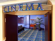 Entrance to an interior cinema room with a large 'CINEMA' sign, rows of seating, patterned carpet, a popcorn cart and potted plants.