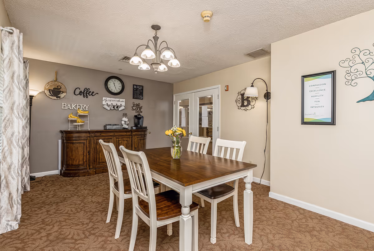 A dining area with a wooden table and four white chairs. A vase with yellow flowers is placed on the table. Behind the table is a wooden sideboard with a coffee station, including a coffee maker, cups, and decorative signs on the wall that say 'Coffee' and 'Bakery'. The room has beige walls, carpeted floor, a chandelier overhead, and a framed sign on the right wall with words like Compassion, Excellence, Humility, Fun, and Integrity.
