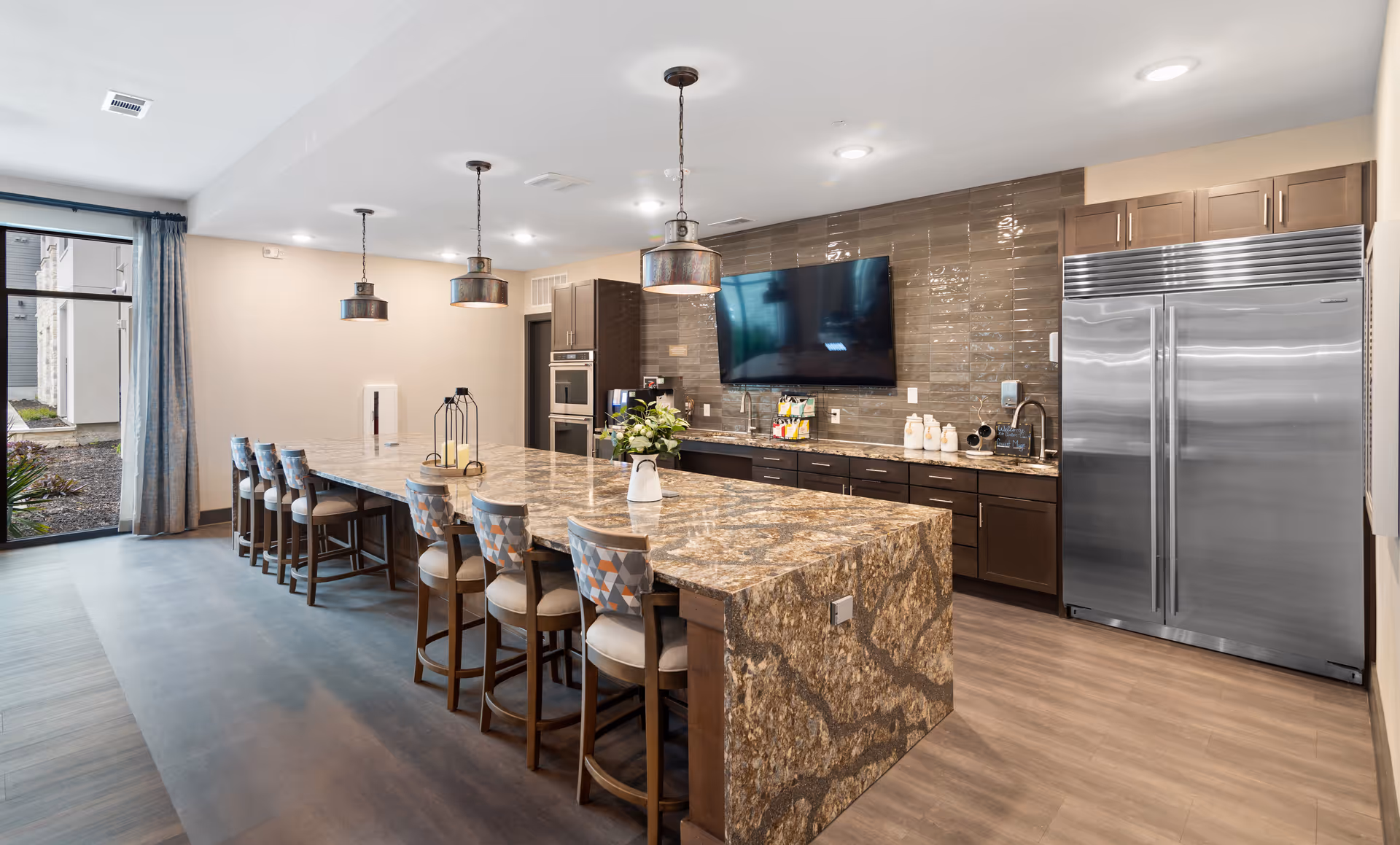 Modern communal kitchen and dining area with a long granite island, bar stools, pendant lights, stainless steel refrigerator and a wall-mounted TV.
