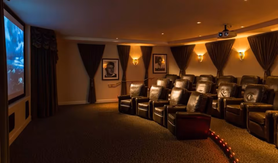 A cozy home theater room with multiple rows of dark leather recliner chairs facing a large screen displaying a sky scene. The room has dim lighting with wall sconces, dark curtains, and framed pictures on the walls.