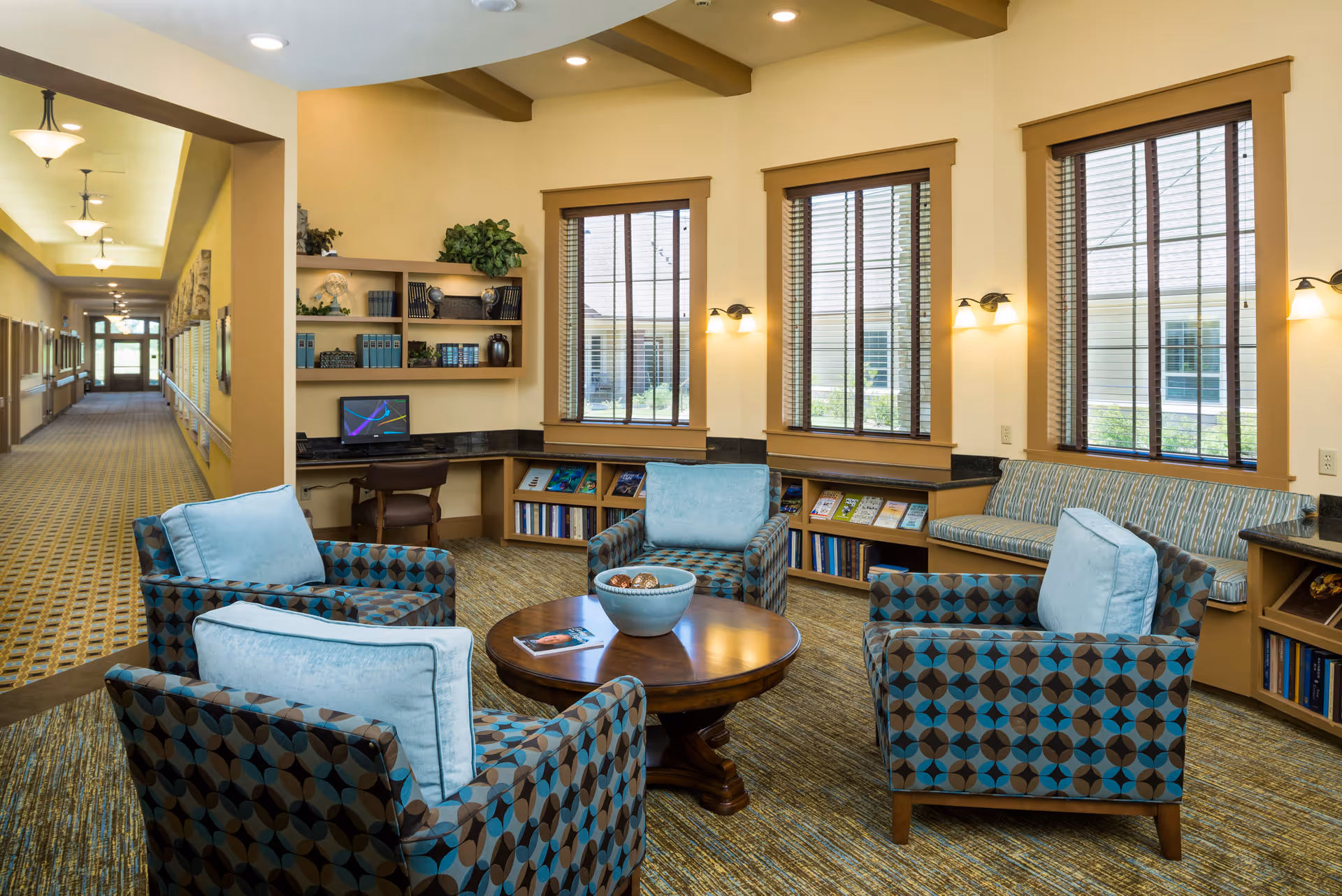 A cozy sitting area in a senior living facility with four patterned armchairs with light blue cushions arranged around a round wooden coffee table. The room has large windows with wooden blinds, built-in bookshelves filled with books and magazines, and a small desk with a computer. The walls are painted beige, and the carpet has a patterned design. A long hallway with ceiling lights extends from the sitting area.
