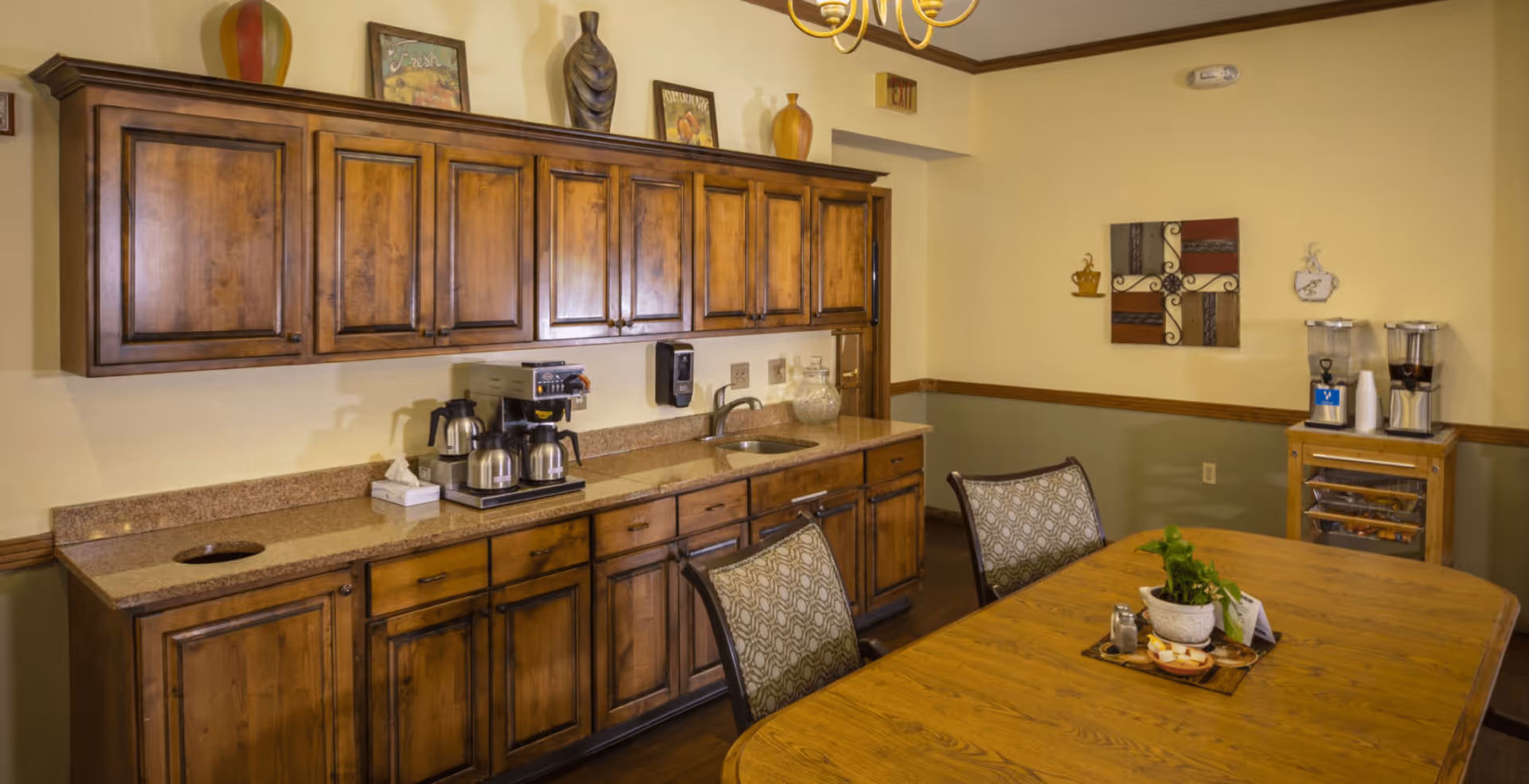 Wooden kitchenette and dining table with chairs, cabinets, a coffee station, and decorative accents in an assisted living dining area.