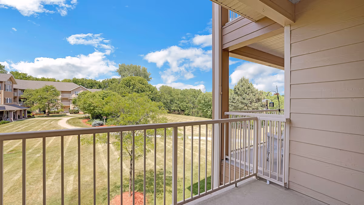 View from a balcony overlooking a grassy area with trees and a walking path, adjacent to a multi-story residential building under a blue sky with scattered clouds.