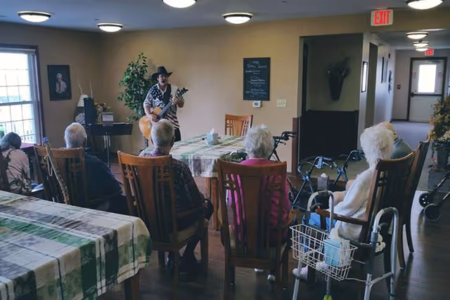 A group of elderly people seated on wooden chairs around tables with checkered tablecloths in a common room, watching a person playing a guitar and singing. The room has beige walls, ceiling lights, and windows letting in natural light. Walkers are visible near some of the seated individuals.