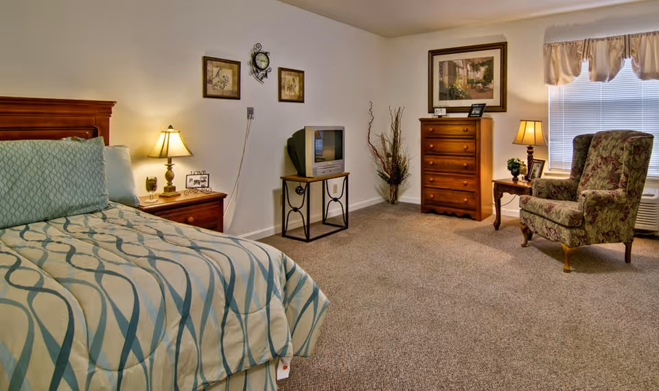 A cozy senior living bedroom featuring a bed with patterned bedding, a wooden nightstand with a lamp and decorative items, a small TV on a stand, a wooden dresser, a floral upholstered armchair, and a window with blinds and a valance.