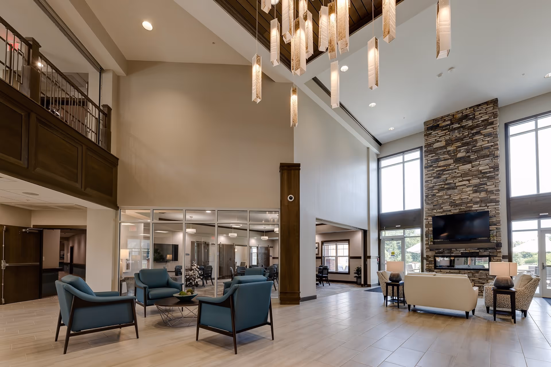Spacious and modern senior living facility common area with high ceilings, a large stone fireplace with a mounted TV, comfortable seating including blue armchairs and a white sofa, large windows letting in natural light, and a contemporary hanging light fixture.