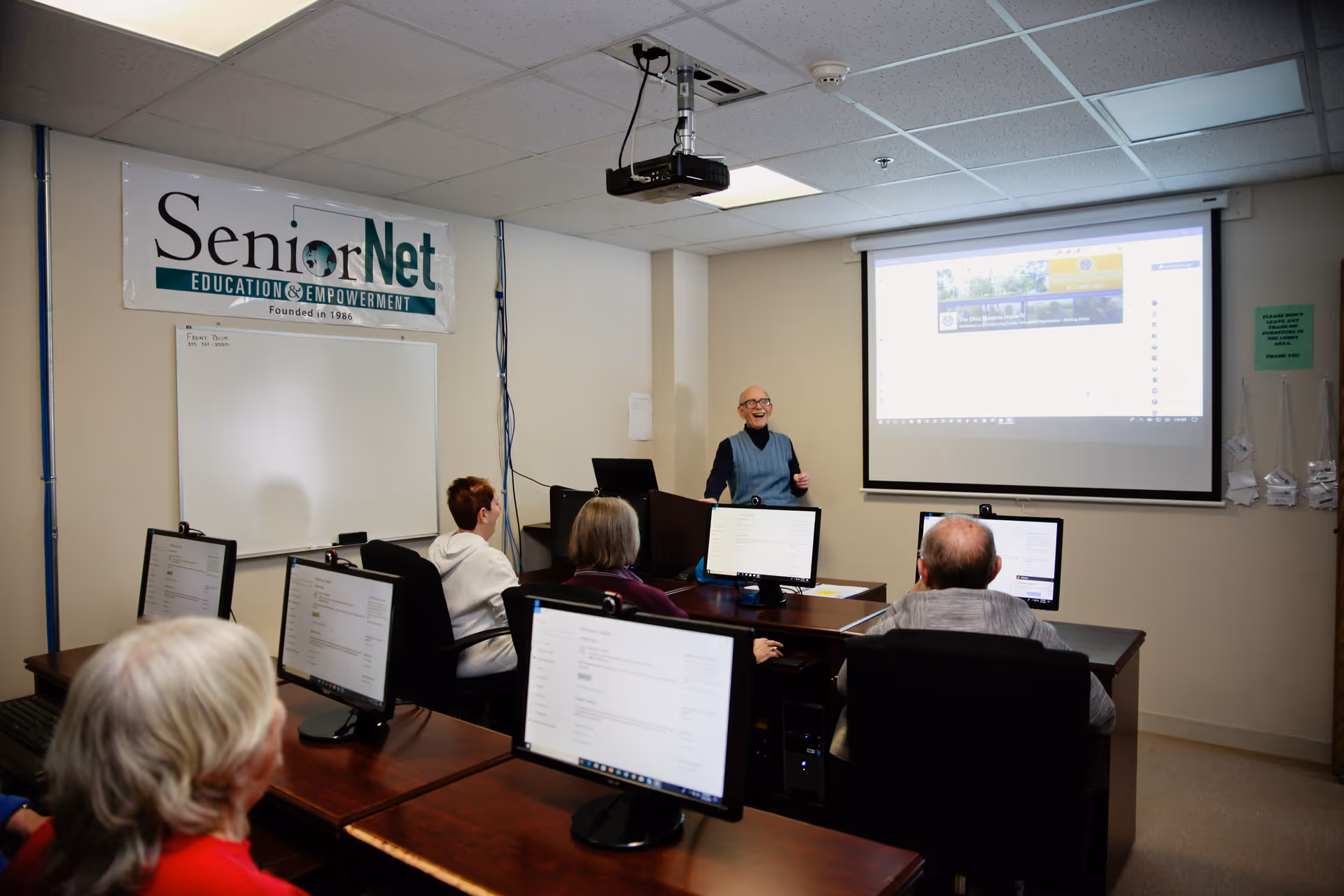 A classroom setting with senior adults seated at desks with computers, facing a woman standing at the front of the room giving a presentation. A projector screen displays a webpage, and a banner on the wall reads 'SeniorNet Education & Empowerment Founded in 1986'.