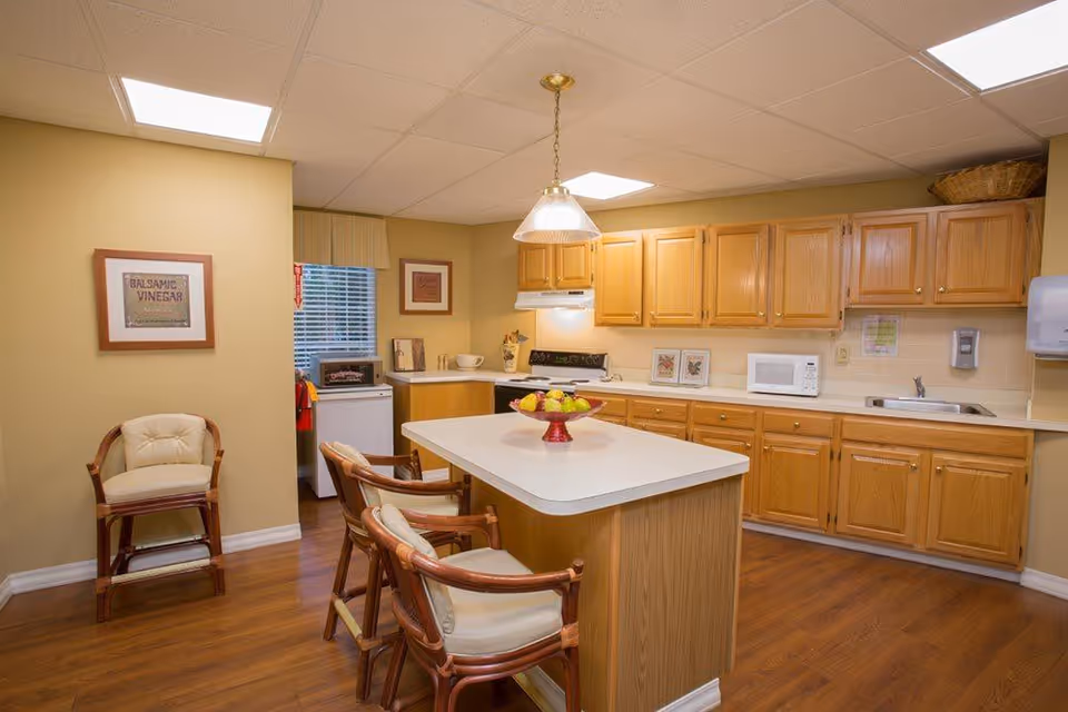 A cozy kitchen area with wooden cabinets, a white countertop island with two cushioned wooden chairs, a bowl of fruit on the island, a microwave, stove, and sink. The room has warm yellow walls, a framed picture on the wall, a window with blinds, and wood flooring.