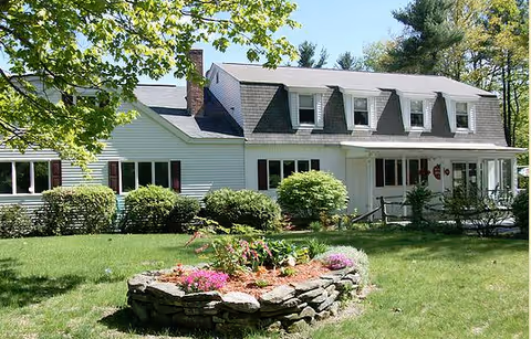 A white two-story house with dormer windows and a front porch, surrounded by green grass, bushes, and trees. In the foreground, there is a circular stone flower bed with blooming pink and purple flowers.