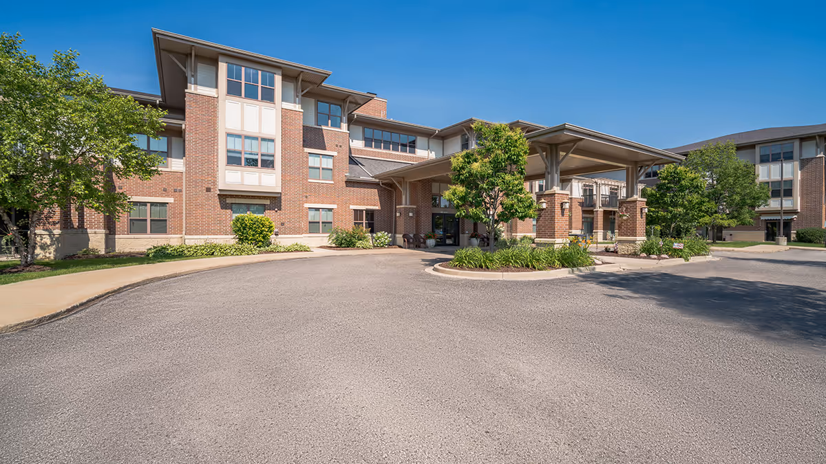 Exterior view of a senior living facility named Village at the Falls, showing a large brick building with multiple windows, a covered entrance with columns, surrounding greenery, and a clear blue sky.