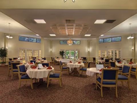 A spacious dining room with multiple round tables covered with white tablecloths and set with red napkins. Each table is surrounded by blue cushioned chairs with wooden frames. The room has a patterned carpet, cream-colored walls, large windows near the ceiling, and multiple light fixtures including a chandelier in the center.