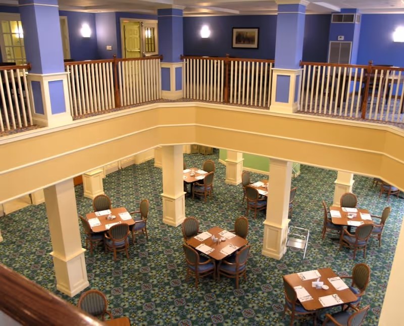 Interior view of a senior living facility dining room with multiple square wooden tables, each set with placemats, napkins, and utensils. The room has a patterned green carpet, cream-colored pillars, and a second-floor balcony with white railings and blue walls. Soft lighting fixtures are mounted on the walls.