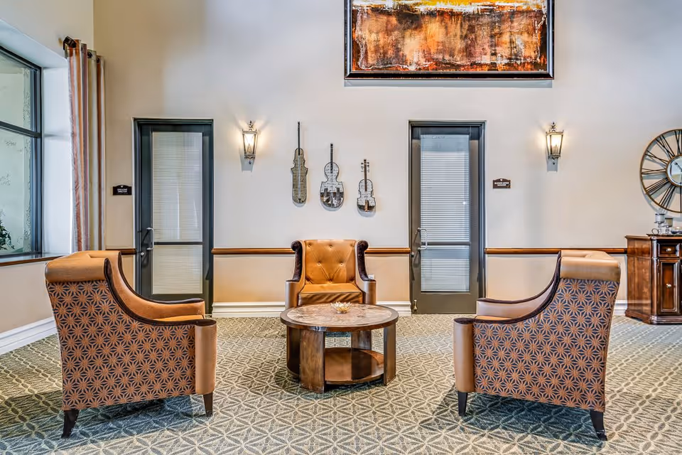 A cozy seating area with three armchairs arranged around a round wooden coffee table. The chairs have a patterned upholstery with brown leather accents. Behind the chairs, there are two glass doors with blinds, wall sconces, and decorative violin-shaped wall art. A large abstract painting hangs above the doors, and a large decorative clock is visible on the right side of the image.