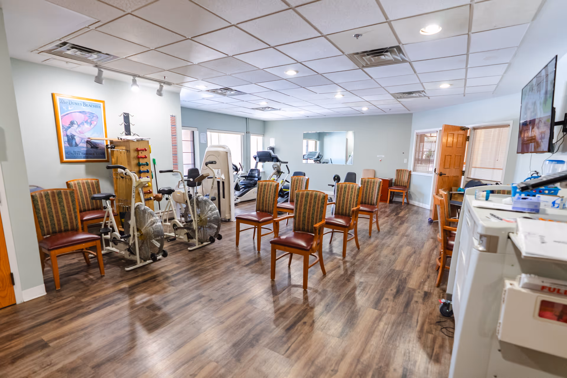 A spacious exercise room with several stationary exercise bikes, chairs arranged in rows, and additional exercise equipment in the background. The room has wood flooring, light-colored walls, a ceiling with recessed lighting, and a large mirror on one wall. There is a door open to another room and a television mounted on the right wall.