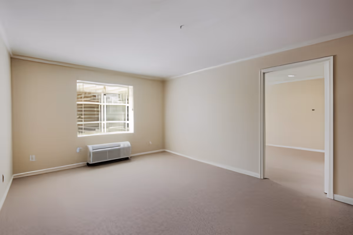Empty carpeted living area with a window, a wall HVAC unit beneath it, and an open doorway to another room.