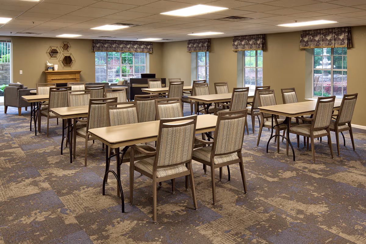 A spacious dining room with multiple rectangular tables and cushioned chairs arranged neatly. The room has large windows with patterned valances, allowing natural light to brighten the space. The carpet has a patterned design, and there is a small seating area with armchairs and a decorative wall shelf in the background.