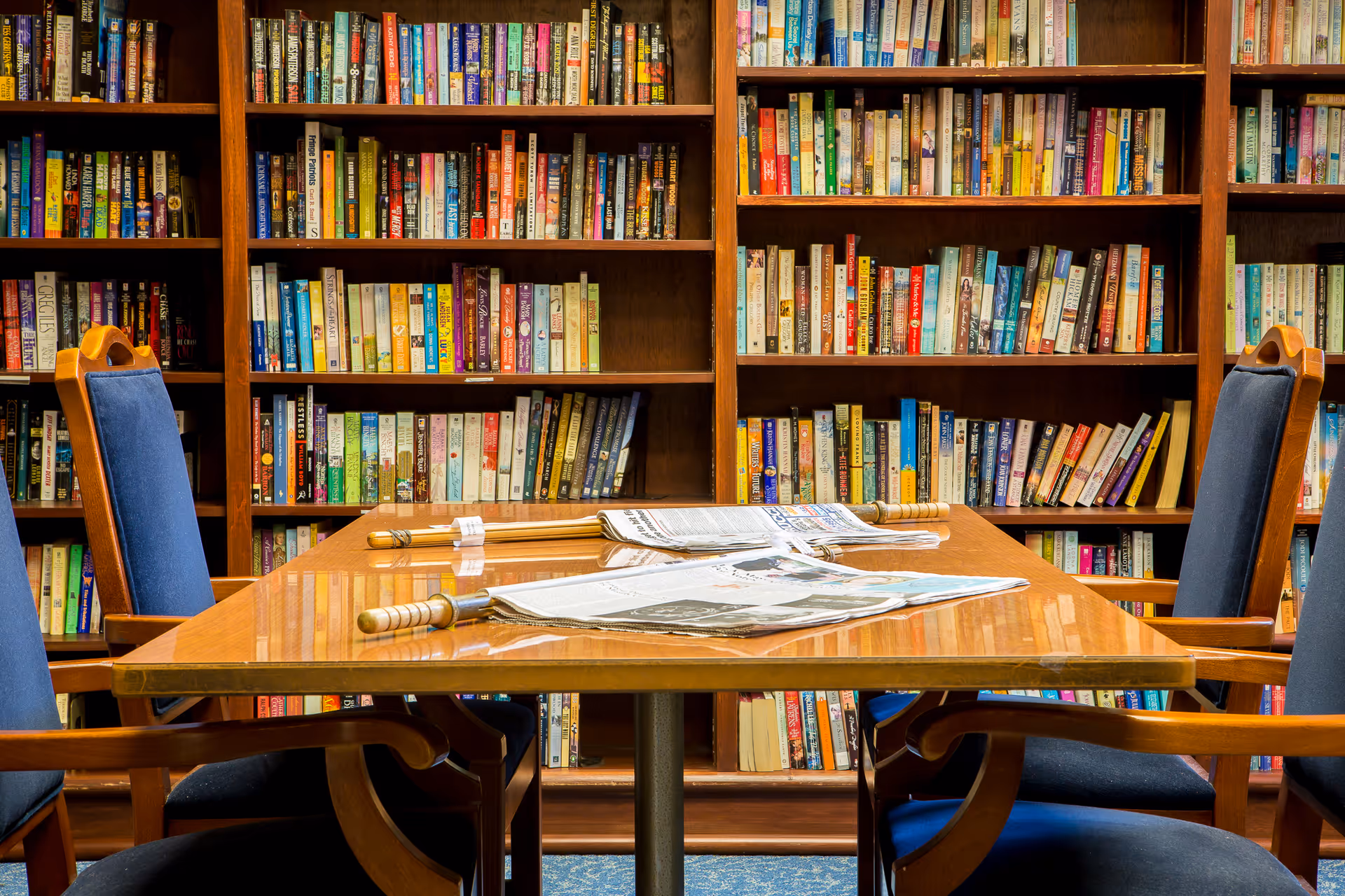 A wooden table with newspapers and wooden-handled magnifying glasses on it, surrounded by blue cushioned wooden chairs, in front of a large bookshelf filled with various books.