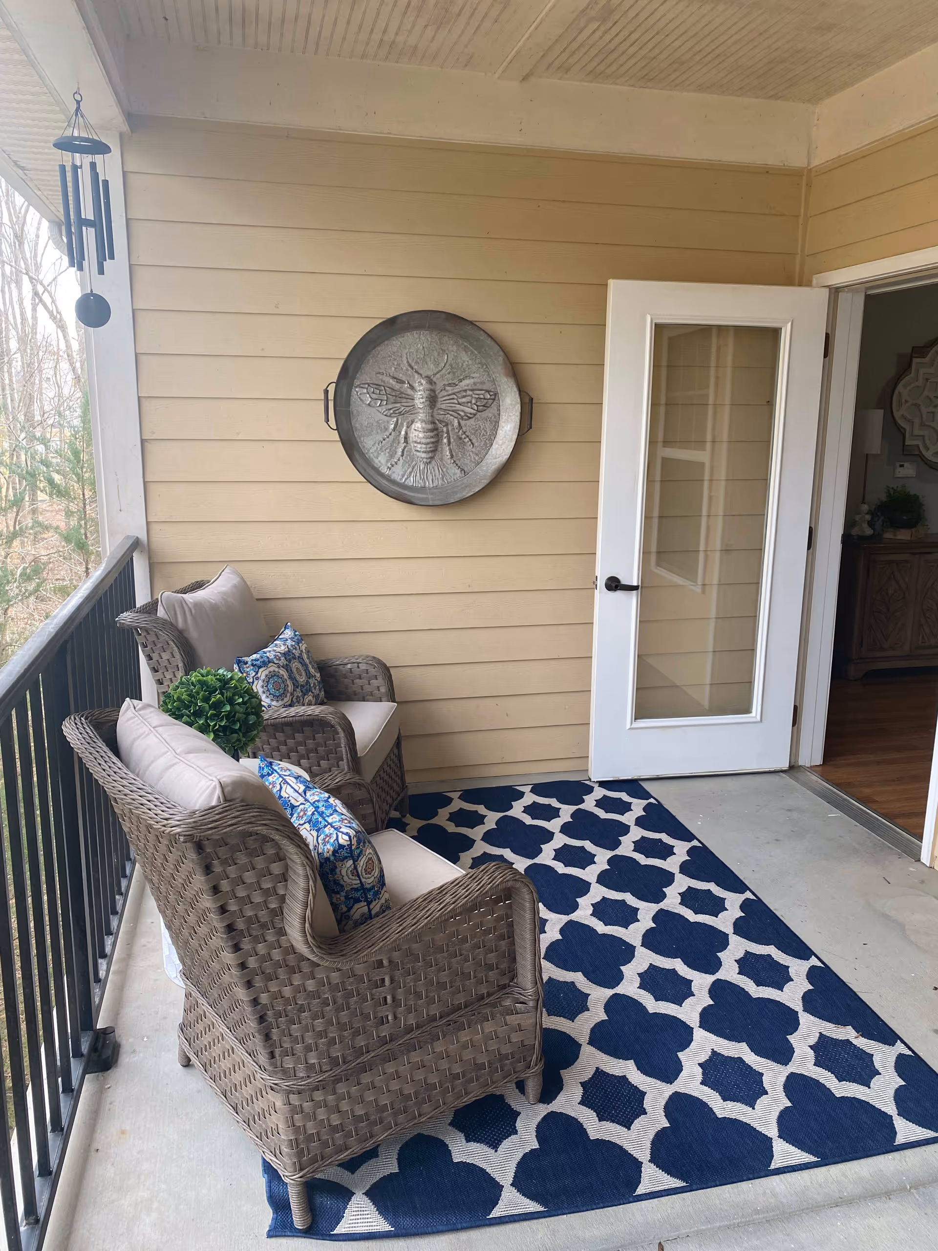 A covered outdoor balcony with two wicker armchairs featuring beige cushions and blue patterned pillows. A blue and white patterned rug covers the floor. A decorative metal wall hanging of a bee is mounted on the beige siding wall. A white door with glass panels is open, leading inside the building. A wind chime hangs from the ceiling on the left side.