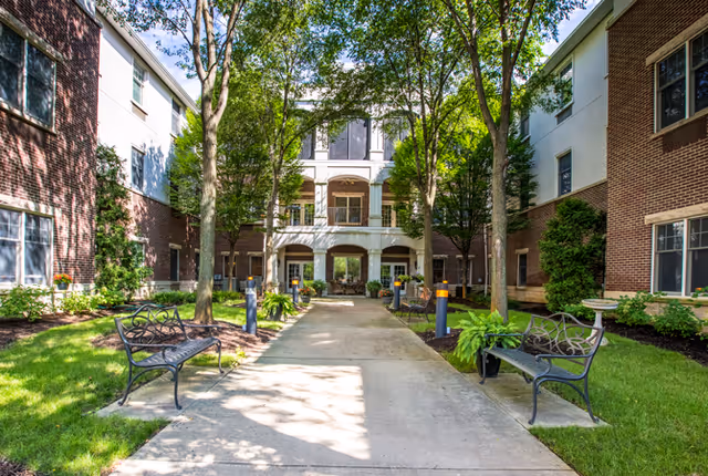Tree-lined courtyard walkway with benches leading to the entrance of a brick senior living building.