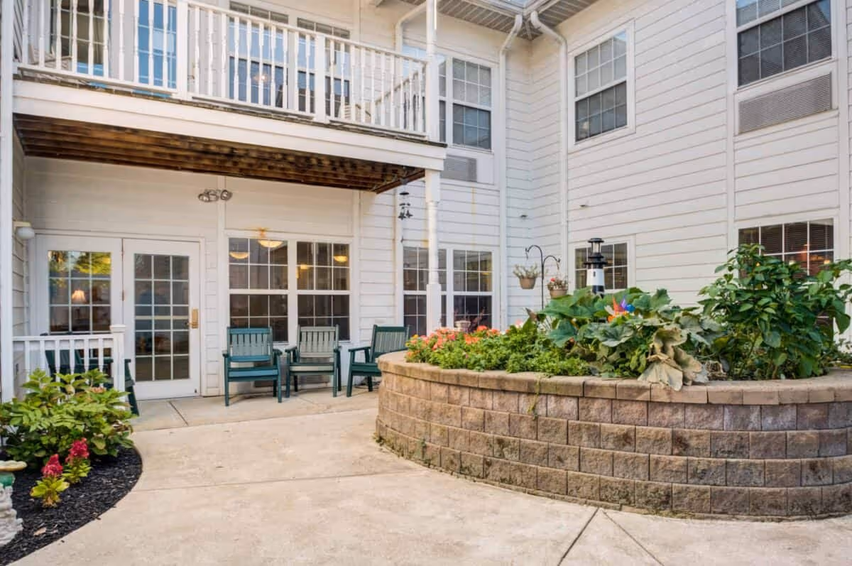 Outdoor courtyard area of a senior living facility with a raised brick planter filled with green plants and flowers. There are several green chairs arranged near white-framed glass doors and windows of the building. The building exterior is white with multiple windows and a balcony above the seating area.